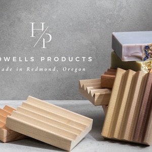 May include: A set of four wooden soap dishes with a variety of designs. The dishes are stacked on top of each other, with a stack of four bars of soap on top. The soap is a variety of colors, including purple, green, and brown. The soap is decorated with dried flowers and herbs. The text "HOWELLS PRODUCTS made in Redmond, Oregon" is visible in the image.