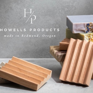 May include: Wooden soap dishes and bars of soap. The soap dishes are made of light-colored wood with a slatted design. The soaps are various colors and textures. The text "HOWELLS PRODUCTS made in Redmond, Oregon" is visible.