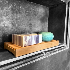 May include: A wooden soap dish holds a rectangular bar of soap with purple and white swirls and a round, light blue bath bomb. The dish is set in a gray tiled shower niche.