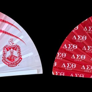 May include: Two red and white folding hand fans with a white Greek letter design and the text "Since 1913". One fan has a floral design on the left side and the other fan has a repeating Greek letter design.