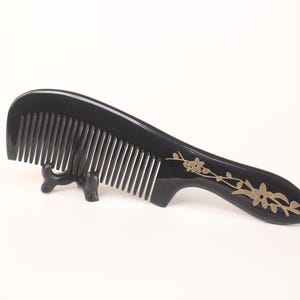 Può includere: Un pettine nero con denti sottili, con un manico curvo ornato da un motivo floreale dorato. Il pettine è esposto su un piccolo supporto nero su sfondo bianco, mostrando il suo design elegante.