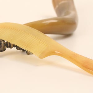 May include: A light yellow comb with a handle rests on a decorative bronze object. The comb has fine teeth and a curved handle. A curved, horn-like object is in the background. The comb is likely used for hair care.