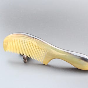 May include: A natural horn comb with a curved handle. The comb has a light yellow color with a gradient of darker hues. The teeth are closely spaced and evenly aligned. This comb is designed for hair care.