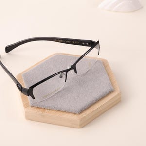 May include: Black rimmed eyeglasses with a semi-rimless design, resting on a wooden hexagonal stand with a gray cleaning cloth. The glasses have a sleek, modern aesthetic. The text "Titanium & Frame" is visible on the lens.