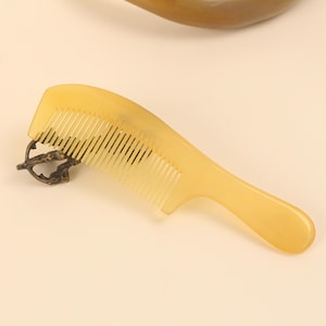 Può includere: Un pettine giallo chiaro con manico e denti fini. Il pettine è realizzato in un materiale liscio e ha un piccolo pezzo di metallo decorativo. Il pettine è progettato per la cura dei capelli.