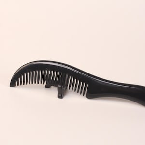 May include: A black, curved hair comb with fine teeth. The comb has a smooth, glossy finish and a unique, ergonomic design. The comb is made of a solid, dark material, likely plastic or a similar substance.