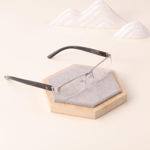 May include: Rimless eyeglasses with black temples and silver frames rest on a wooden hexagonal display stand with a gray fabric insert. The glasses are positioned in front of a white, abstract mountain-like sculpture.