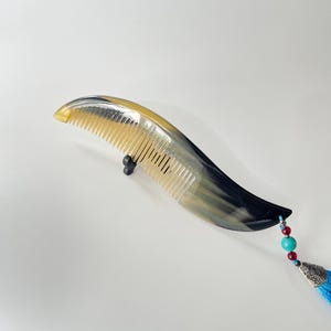 Può includere: Un pettine curvo per capelli realizzato in corno lucido con un motivo sfumato giallo e nero traslucido. Il pettine presenta un attacco decorativo con piccole perline rosse e turchesi, un ciondolo in metallo argentato e una nappa blu brillante.