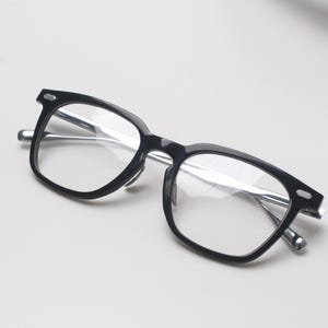 May include: Black-framed eyeglasses with clear lenses. The glasses have a rectangular shape with rounded edges and silver-colored temples. The frames are made of a glossy black material.