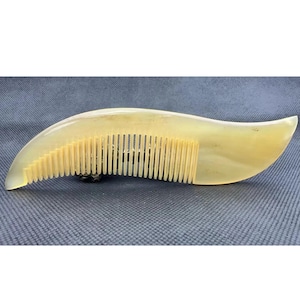 Puede incluir: Un peine amarillo claro con un mango curvo y dientes finos. El peine tiene un acabado liso y pulido y una apariencia natural y orgánica. El peine está diseñado para el cuidado y peinado del cabello.