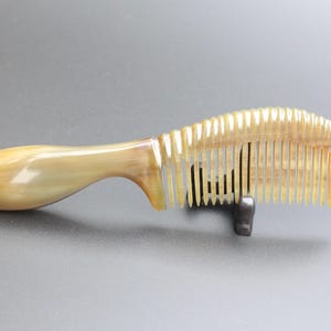 May include: A natural horn comb with a curved handle and closely spaced teeth. The comb is a light tan color with a glossy finish. The handle curves smoothly into the comb's teeth. The comb is designed for hair care.