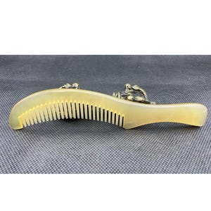 May include: A curved, light-colored comb with fine teeth. The comb features decorative metal embellishments near the handle. The comb is made of a light-colored material, possibly horn or bone, and is designed for hair care.