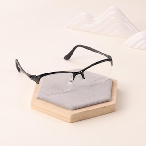 May include: Black-framed eyeglasses with a half-rim design, resting on a wooden hexagonal stand with a gray cleaning cloth. The glasses have clear lenses. A white, abstract mountain-shaped decoration is in the background.