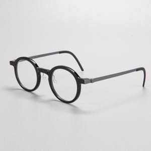 May include: A pair of round-framed eyeglasses. The frames are black with gray temples. The lenses are clear. The glasses are set against a light gray background.