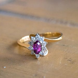 以下が含まれることがあります： 中央にマーキスカットのマゼンタ色の宝石を配し、その周囲をホワイトメタルの台座にセットされた小さな透明なラウンドカットの石で囲んだゴールドリング。リング部分は滑らかで光沢のあるイエローゴールドで、中央に向かって細くなっています。