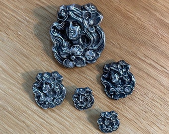 May include: A set of five silver-toned metal buttons featuring a woman's face with floral accents. The largest button is a pendant with a detailed design, while the smaller buttons are round and have a simpler design.