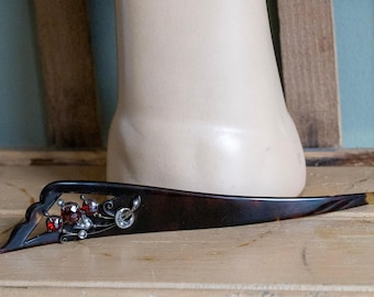 May include: A dark brown, tapered hair stick featuring an openwork design at the wider end. This section is embellished with three faceted red stones and small, clear, round accents set within a silver-toned metal frame with delicate swirls.