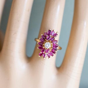 May include: A gold ring featuring a central round white gemstone surrounded by two concentric layers of marquise-cut deep pink rubies arranged in a floral cluster pattern. The setting is crafted from yellow gold, showcasing a vibrant contrast between the central stone and the surrounding pink petals.