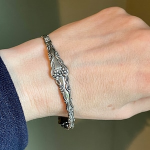 May include: A silver bracelet made from a vintage silverware piece. The bracelet has a floral design and is worn on a person's wrist.