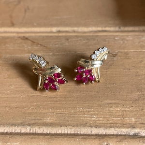 May include: A pair of gold-toned earrings featuring a cluster of small, marquise-cut, ruby-colored gemstones and a row of clear, round-cut gemstones. The earrings have a stylized, bow-like design.