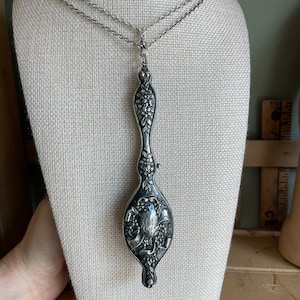 Op de afbeelding: Een zilveren ketting met een gedetailleerd bloemmotief hanger. De hanger is gevormd als een lange, dunne traan met een bloemmotief en een klein, rond, gegraveerd ontwerp aan de onderkant.
