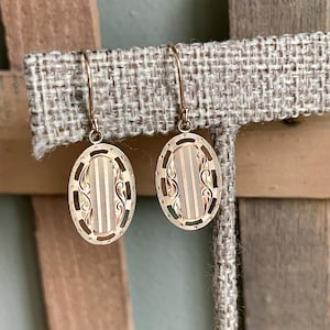 May include: A pair of gold-colored oval earrings with a vertical striped design and decorative scrollwork. The earrings are suspended from a simple hook. The earrings are displayed on a neutral-colored fabric.