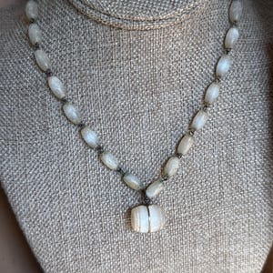 Pode incluir: Um colar vintage com contas ovais brancas sujas, unidas por uma corrente de metal escuro. Apresenta um pingente retangular com uma faixa de metal prateado. O colar é exibido em um manequim de cor neutra.