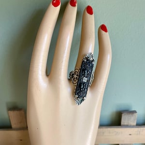 May include: An Art Deco ring featuring a black onyx centre stone and two small diamonds, set in silver. The ring has intricate filigree detailing and is displayed on a mannequin hand with red nail polish.