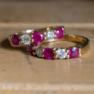 Könnte beinhalten: Zwei goldene Ringe mit abwechselnd roten Edelsteinen und klaren Diamanten. Die roten Steine wechseln sich mit den Diamanten ab. Die Ringe liegen auf einer hellbraunen Holzoberfläche, die ihren Glanz hervorhebt. Ideal für Verlobung oder Hochzeit.
