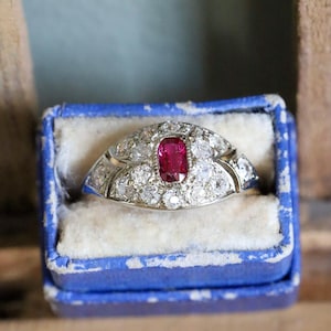 Könnte beinhalten: Ein silberfarbener Ring mit einem zentralen, oval geschliffenen roten Edelstein, der von einer Gruppe runder, klarer Steine umgeben ist. Der Ring hat eine gewölbte Fassung mit mehreren Steinen und eine polierte Metallschiene in einer blauen Box.