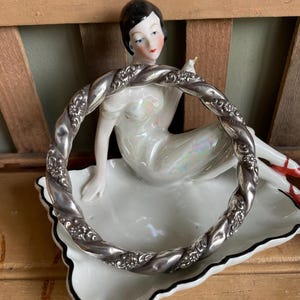 Puede incluir: Un brazalete de plata trenzado con detalles florales. El brazalete se muestra en un plato de porcelana blanca con un borde negro.