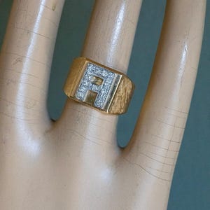 May include: A gold signet ring featuring a rectangular face with a central letter A inlaid with small clear stones. The band has a textured finish on the sides, and the metal has a polished gold tone throughout the main body of the ring.