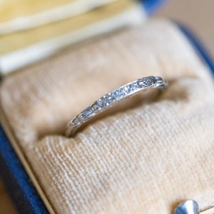 May include: A thin silver-toned metal band ring featuring a series of small, clear, sparkling stones embedded along the top half of the band. The ring is set into a soft, beige, velvet-lined interior of a small, dark blue, hinged jewelry box.