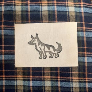 May include: An off-white fabric patch with a grayscale illustration of a dog. The dog is in a standing pose with a curled tail. The patch is set against a plaid fabric background in shades of blue, green, and orange.