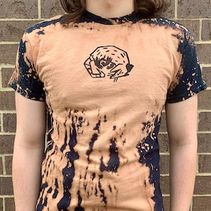 Puede incluir: Una camiseta de color marrón claro con un gráfico en blanco y negro de un cangrejo en la parte delantera. La camiseta tiene un diseño salpicado de lejía en negro.