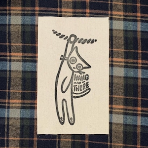 May include: A black and white patch with a cartoon character hanging from a rope. The character has a large nose and holds a sign saying "Hang in There". The background is a plaid pattern.