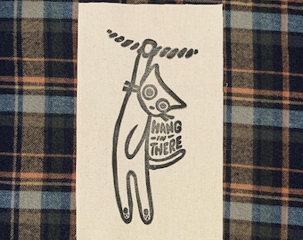 May include: A black and white patch featuring a cartoon figure hanging from a rope. The figure has a large nose and holds a sign that reads "Hang in There". The background is a plaid pattern.