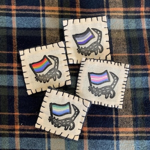 May include: Four square patches with a cream-colored background and black stitching. Each patch features a different LGBTQ+ pride flag design with a black cat silhouette. The flags include rainbow, asexual, transgender, and non-binary pride flags.