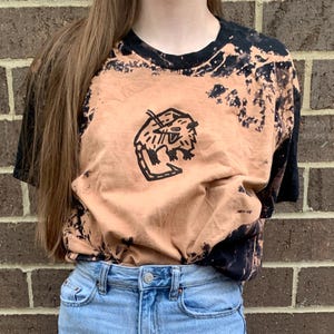 May include: A light brown t-shirt with a black and white graphic of a cartoon animal. The shirt has a bleach-splattered design.