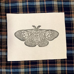 May include: A grey moth illustration with detailed wing patterns is printed on a rectangular piece of off-white fabric. The moth design is centred and the fabric is placed on a plaid background.