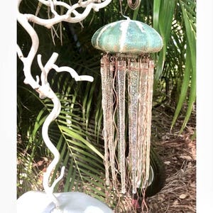 May include: A teal and white ceramic jellyfish wind chime with clear glass beads hanging from copper wire. The chime is hanging from a white branch.