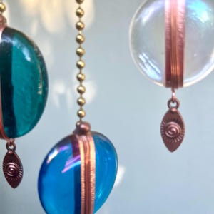 May include: Three glass ornaments with copper accents hang from gold chains. The ornaments are blue, green, and clear. Each ornament has a copper charm hanging from the bottom.