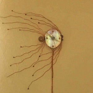 May include: A wall clock featuring a circular iridescent mother-of-pearl face with dark hands. Thin, brown wire tendrils extend outward from the left side of the clock, each tipped with a small dark bead, creating a radiating, organic, fan-like pattern.