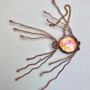 May include: A copper wire ornament with a round iridescent glass center and a gold chain. The ornament has multiple copper wires that extend from the center and curve outward.