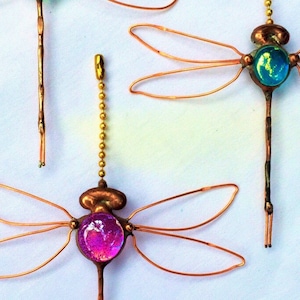 May include: Three copper wire dragonfly ornaments with glass accents. The dragonflies have iridescent wings and a variety of colored glass eyes. The dragonflies are hanging from a chain.