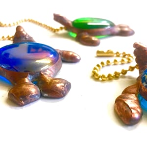May include: Three handcrafted turtle pendants with iridescent blue and green glass shells and copper-colored metal bodies. Each turtle is attached to a gold-colored ball chain. The pendants are displayed on a white surface.