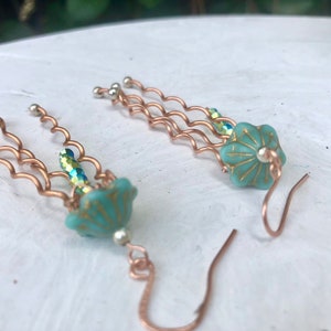 May include: A pair of dangle earrings with a turquoise blue and gold flower charm. The earrings are made with copper wire and have a wavy design. The earrings are approximately 2 inches long.