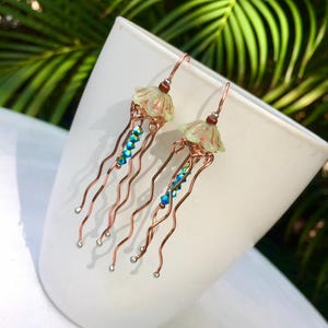 May include: A pair of copper wire earrings with a light green glass jellyfish charm and blue crystal beads hanging from each. The earrings are dangling and have a delicate, whimsical look.