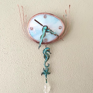 May include: An oval wall clock with a white iridescent glass face, copper wire frame, and wavy turquoise hands. A dangling ornament features a turquoise seahorse, a starfish, and a clear crystal pendant hanging from the bottom of the clock.