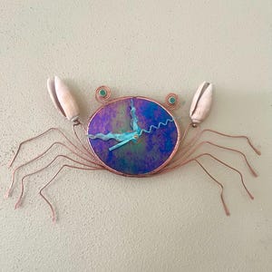May include: A crab-shaped wall clock with copper wire legs and claws. The clock face is a circular iridescent glass with blue and purple hues. The crab's eyes are small green beads, and the claws are made of white seashells.